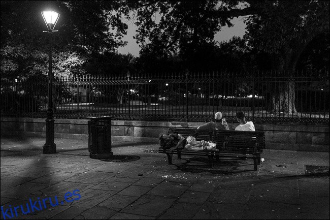 dos hombres sentados en un banco del parque por la noche;  un hombre durmiendo en el asiento detrás de ellos