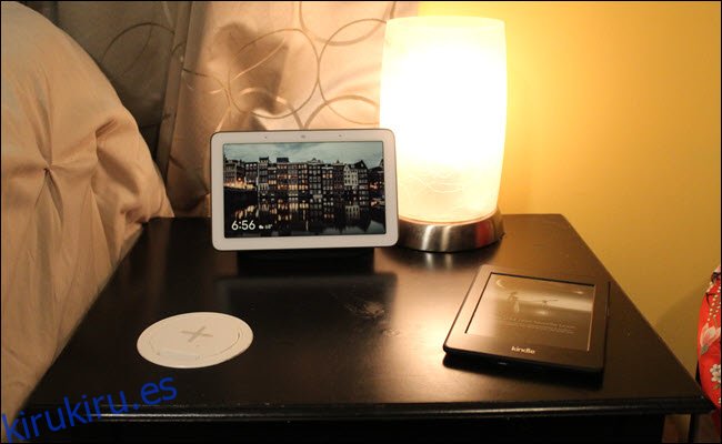 Una mesita de noche con un cargador inalámbrico instalado en la superficie, un Kindle y Google Home Hub