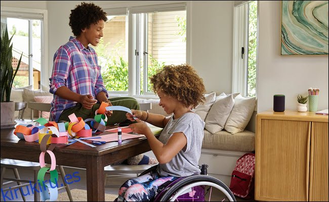 Una niña pequeña en silla de ruedas haciendo manualidades con su mamá en una mesa con un Amazon Echo cerca.