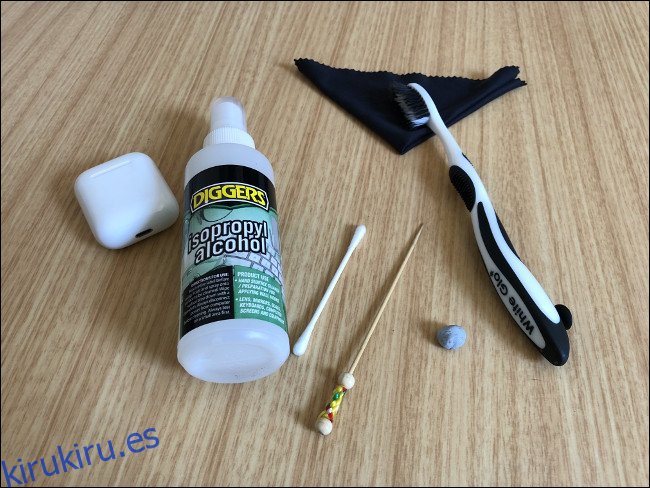 Un estuche de carga de AirPods sobre una mesa junto a una botella de spray de alcohol isopropílico, un hisopo, un palillo de dientes, Blu-tack, un cepillo de dientes y un paño. 