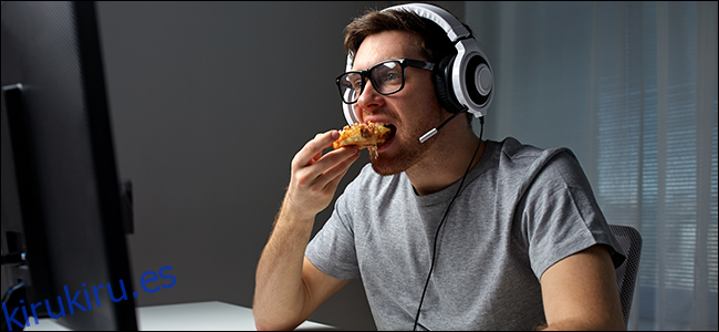 Un hombre sentado frente a una computadora, con auriculares y comiendo pizza.